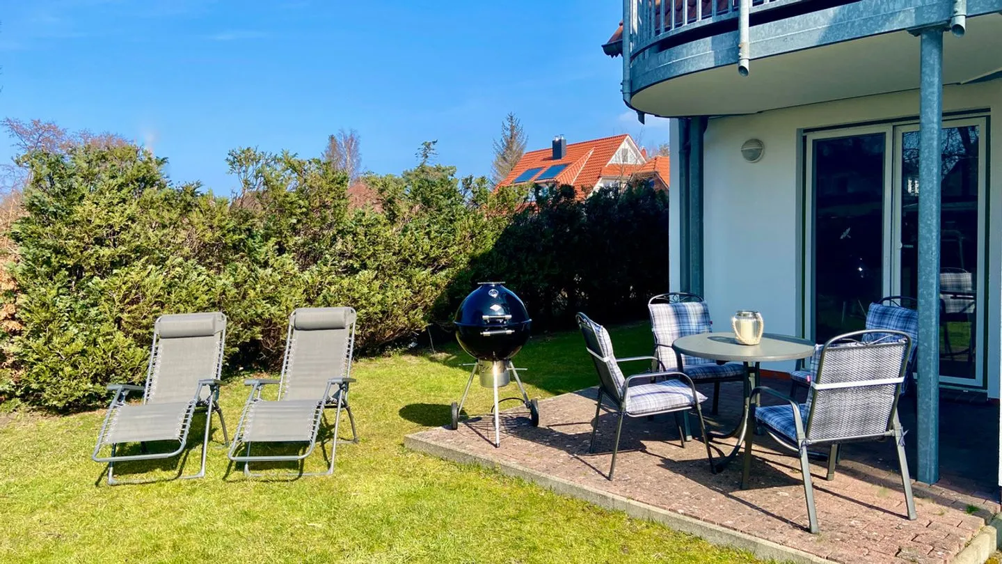 Sonnige Terrasse mit Sitzgruppe, Kugelgrill und zwei Gartenliegen auf dem Rasen vor dem Haus